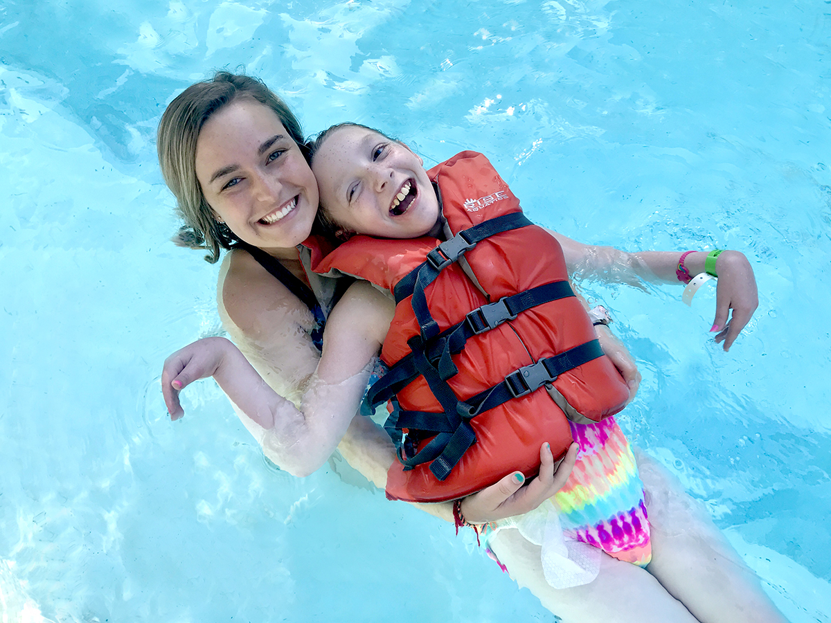 Counselor and camper smiling happily as they float in the pool with life jackets on. 