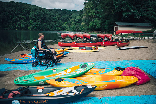 A young adult in a wheelchair navigates among kayaks at the beach on new accessibility mats. 