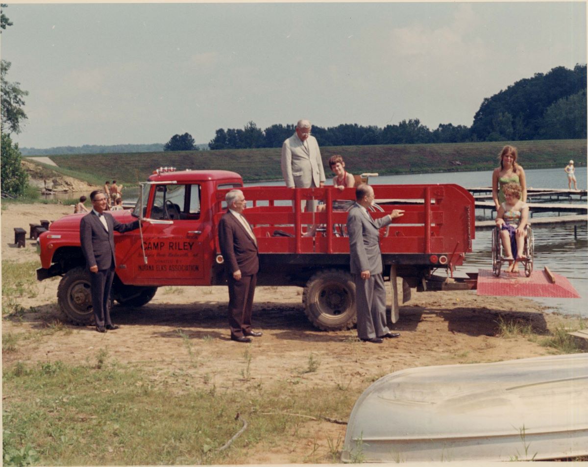 Campers using wheelchairs are loading and unloading from a pickup truck that says "Camp Riley" at the beach. The photo appears to be from the 1960s.