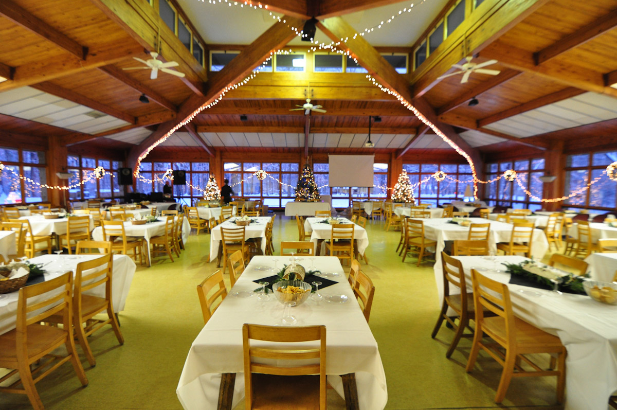 The inside of a large dining hall, tables set with white linens. Christmas lights are hung along the outside walls.