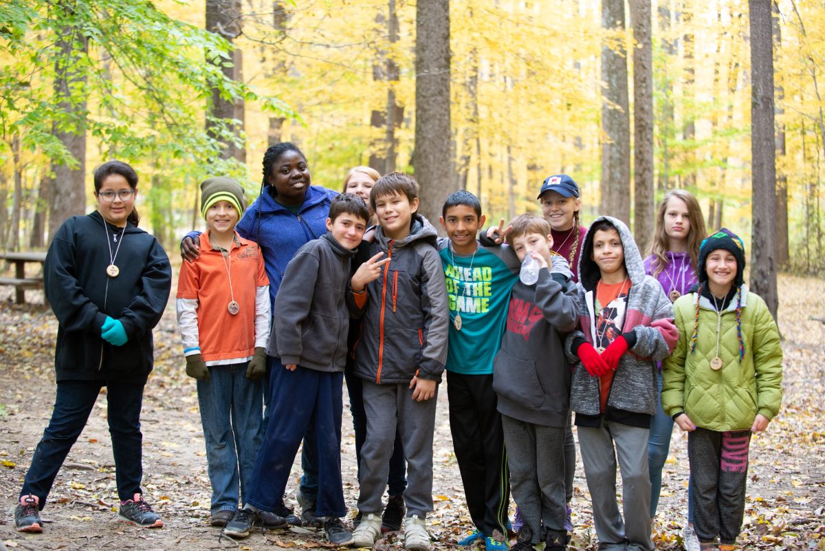 A smiling group of students with their outdoor instructor in front of a canopy of yellow autumn leaves.